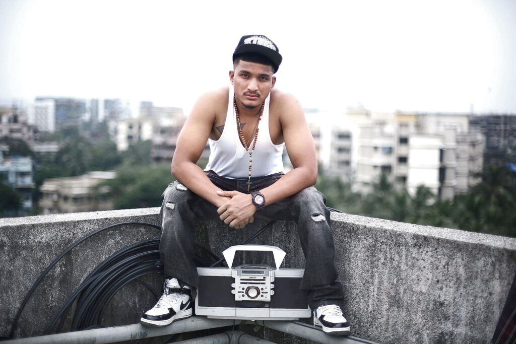 Young Indian rapper Divine in his early days, sitting on a rooftop with a boombox, wearing a cap, white tank top, and ripped jeans, with an urban cityscape in the background